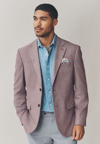 Lys lilla, struktureret blazer over en lyseblå skjorte med knapper, med et mønstret lommetørklæde. Grå bukser fuldender outfittet.