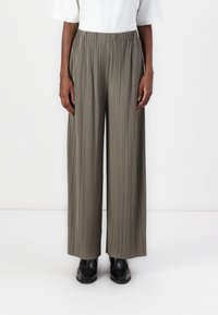 Personne portant un pantalon large à taille haute, plissé et de couleur vert olive, associé à des bottes noires à bout pointu, se tenant contre un fond blanc.