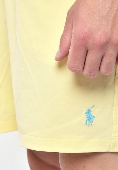 Pantaloni corti gialli realizzati in tessuto leggero, con un logo blu ricamato di un giocatore di polo sul lato sinistro. Texture liscia, lunghezza al ginocchio.
