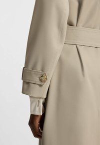 Beiger Trenchcoat mit tailliertem Bund, ausgestattet mit einem Knopfverschluss an den Ärmeln und einer glatten, satinähnlichen Fütterung. Schlichtes Design mit klaren Linien und taillierter Passform.