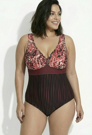 Mujer con cabello oscuro hasta los hombros sonríe llevando un bañador de una pieza sin mangas con parte superior floral y parte inferior a rayas en burdeos y negro.