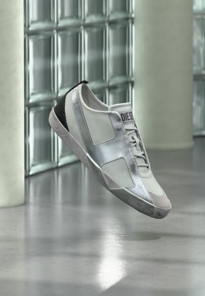 Zapatilla blanca y plateada de la marca Diesel flotando sobre un suelo reflectante con pared de bloques de vidrio y columnas al fondo.