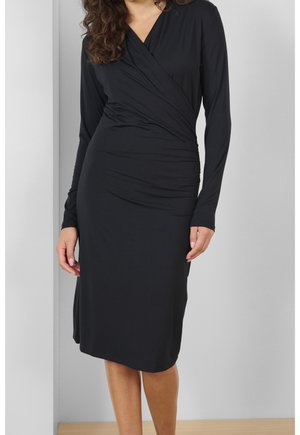 Kobieta ubrana w czarną, długorękawową sukienkę kopertową z marszczeniami po jednej stronie, stojąca na tle jasnego, jednolitego tła.