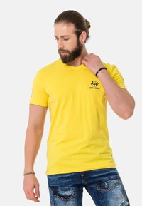 Gelbes V-Ausschnitt-T-Shirt aus Baumwolle mit einem kleinen schwarzen Logo auf der Brust. Der Stoff ist glatt und hat ein tailliertes Design.