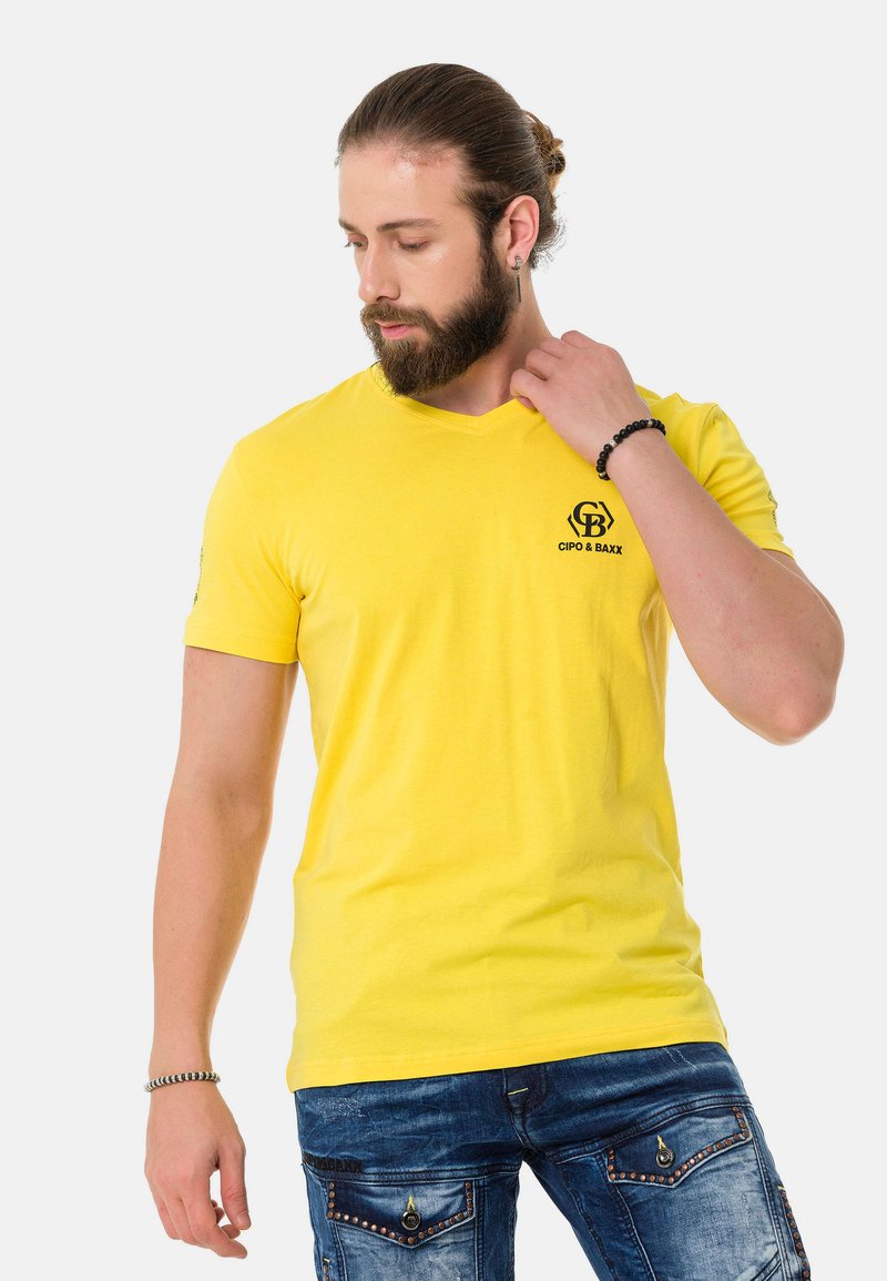 Gelbes V-Ausschnitt-T-Shirt aus Baumwolle mit einem kleinen schwarzen Logo auf der Brust. Der Stoff ist glatt und hat ein tailliertes Design.