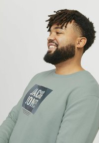 Sudadera verde claro con cuello redondo, que presenta un estampado gráfico en azul y blanco, mostrando "JACK JONES" en la parte delantera.