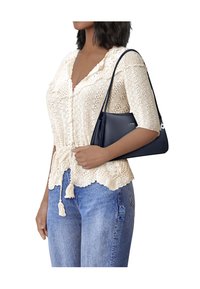 Vrouw draagt een crèmekleurig gehaakt vest en blauwe spijkerbroek, met een marineblauwe leren schoudertas met korte riem aan haar arm.