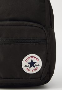 Zaino nero realizzato in tessuto resistente, dotato di una tasca frontale con zip e una toppa circolare con un'icona a stella e testo.