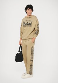 Zestaw z odzieżą w kolorze brązowym, składający się z bluzy z kapturem i spodni, z czarnymi nadrukami graficznymi "Aries", elastycznymi mankietami i kapturem ze sznurkiem. Uzupełniony białymi sneakersami i czarną torbą.