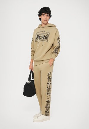 Conjunto de moletom e calças em tom areia com impressões gráficas pretas de "Aries", punhos elásticos e capuz com cordão. Usado com ténis brancos e uma bolsa preta.