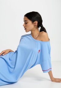 Gaiši zilā krāsā oversized t-krekls, kas izgatavots no mīksta auduma, ar plašu kakla izgriezumu, īsām piedurknēm un sarkanu logo uz krūtīm.