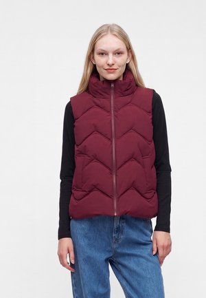 Femme aux longs cheveux blonds portant un gilet matelassé sans manches bordeaux sur un haut noir à manches longues et un jean bleu, debout sur un fond uni.