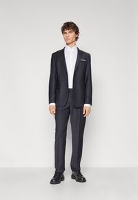 Dark gray tailored suit with notch lapels, single button closure, and a white dress shirt. Completing the look are black lace-up shoes.