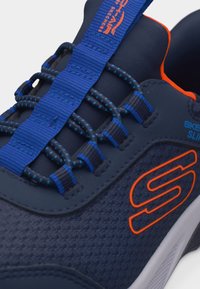 Σκουρό μπλε αθλητικό παπούτσι Skechers με πορτοκαλί λογότυπο, μπλε ελαστικά κορδόνια και μπλε ταινία έλξης με πορτοκαλί κείμενο στη γλώσσα.