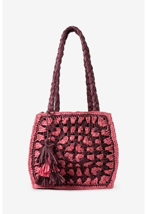 Sac cabas carré tissé à la main avec poignées tressées, présentant un fil épais rose et violet foncé et des décorations à pompons sur fond blanc.