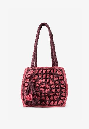Sac cabas carré tissé à la main avec poignées tressées, présentant un fil épais rose et violet foncé et des décorations à pompons sur fond blanc.