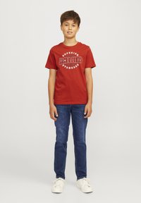 Raudona medvilninė marškinė su balta užrašu "SUPERIOR JACK & JONES STANDARD" priekyje. Derinama su tamsiai mėlynomis džinsinėmis kelnėmis ir baltais sportbačiais.