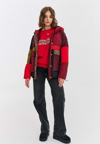 Mladá žena oblečená v červenom grafickom svetri, pufovej bunde z patchworku s leopardím lemom, čiernych džínsoch a čiernych platformových topánkach, stojí na bielom pozadí.