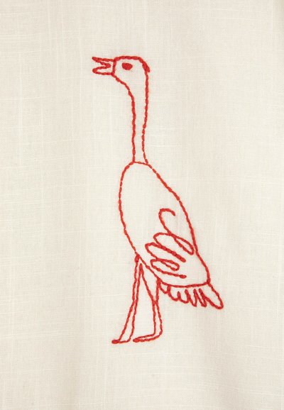 Broderie rouge d'un oiseau debout au bec ouvert avec des ailes détaillées, cousue sur un fond de tissu blanc cassé.