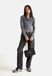 Ung kvinde iført en grå zip-up jakke og sorte jeans, som holder en sort quilted håndtaske med guldkæder foran en hvid baggrund.