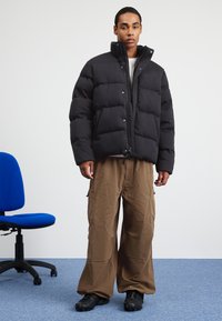 Jachetă puffer neagră cu guler înalt și cusături vizibile, purtată cu pantaloni cargo maro largi și pantofi negri, stând pe un covor albastru.