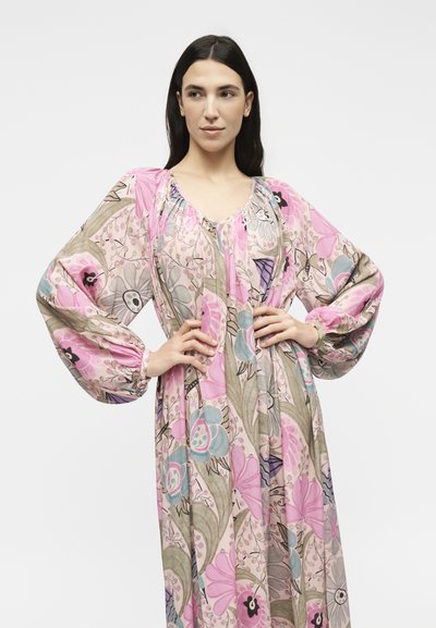 Γυναίκα με μακρύ φόρεμα με λουλούδια σε ροζ, μοβ και πράσινα σχέδια, στέκεται με τα χέρια στη μέση μπροστά σε απλό φόντο.