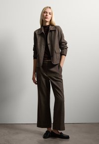 Veste en mélange de laine marron avec un col, des poches avant et une fermeture à boutons ; associé avec un pantalon large assorti et des mocassins noirs.