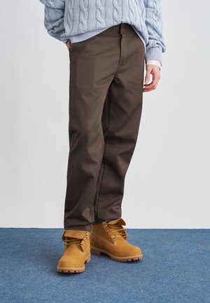 Person trägt braune, gerade geschnittene Hose, beige Schnürstiefel und einen hellblauen Zopfpullover und steht auf einem blauen Teppich.