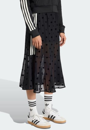 Osoba nosząca czarną siateczkową spódnicę w groszki, czarną kurtkę z białymi paskami, białe skarpetki Adidas oraz białe buty sportowe Adidas z czarnymi paskami.