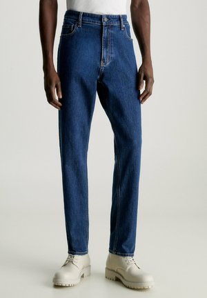 Jeans in denim blu scuro con taglio dritto, cinque tasche e cuciture sottili. Indossati con scarpe con suola spessa di colore chiaro.