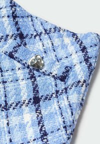 Tissu en tweed bleu avec un motif à carreaux, doté d'accents blancs et bleu foncé. Un bouton triangulaire en argent est visible sur le revers.