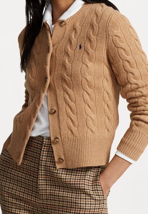 Mujer con cárdigan de punto grueso color camello sobre camisa blanca y pantalones con estampado de pata de gallo, con la mano en el bolsillo.