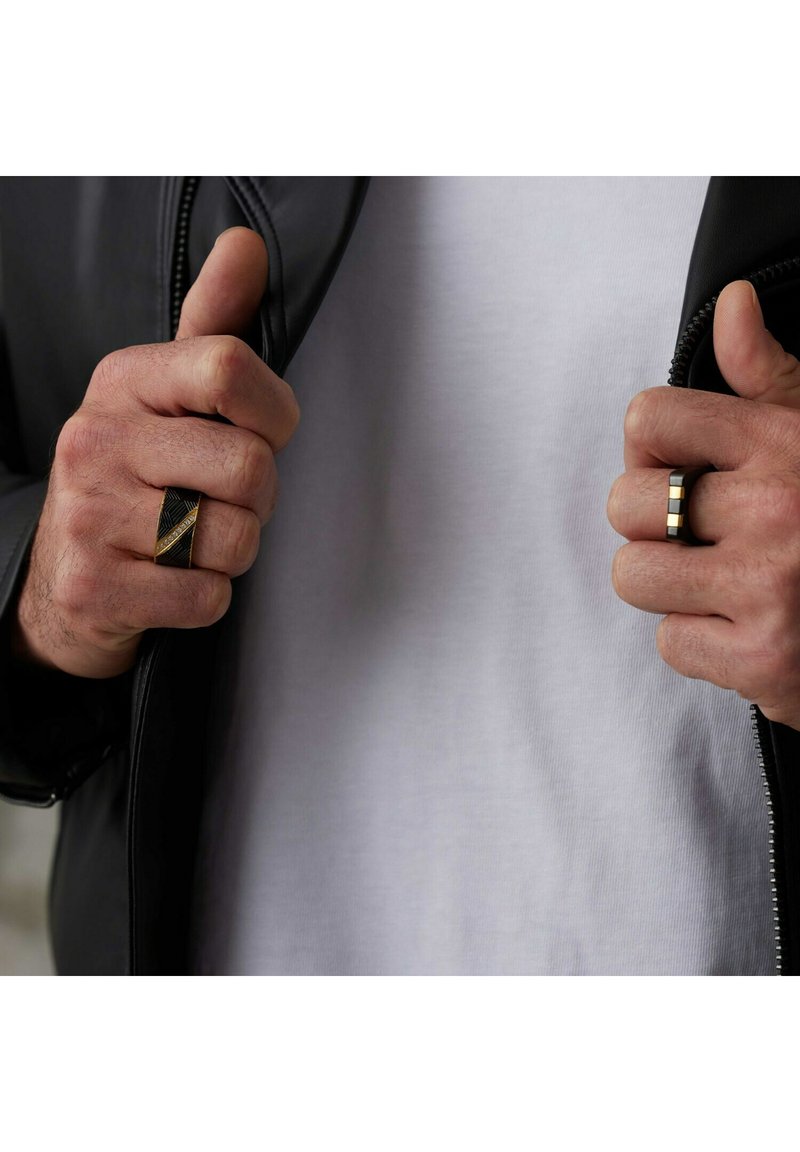 Twee ringen worden gedragen aan de handen: een zwarte ring met een gouden streep, de andere een zwarte band met twee gouden accenten. De achtergrond heeft een zwart jasje en een witte shirt.