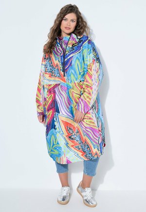 Žena oblečená v barevném, přehnaně velkém kabátu s abstraktními vzory, krátkých džínách a stříbrných teniskách, stojící na bílém pozadí.