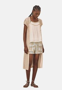 Un cárdigan largo de ganchillo beige ligero sobre una camiseta blanca, combinado con pantalones cortos cargo azul claro estampados y sandalias marrones con tiras.