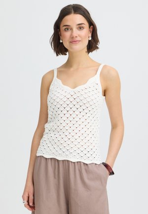 Nő, aki fehér kötött ujjatlan felsőt és bő bézs nadrágot visel, sima világos háttér előtt pózol.