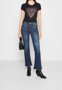 Camiseta de algodón negra con un logo triangular rojo, combinada con unos vaqueros de mezclilla azul de campana y botines negros con tacón, sosteniendo un bolso negro.