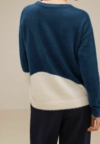 Person som bär en blå och vit fluffig tröja med långa ärmar, sedd bakifrån, mot en enkel beige bakgrund.