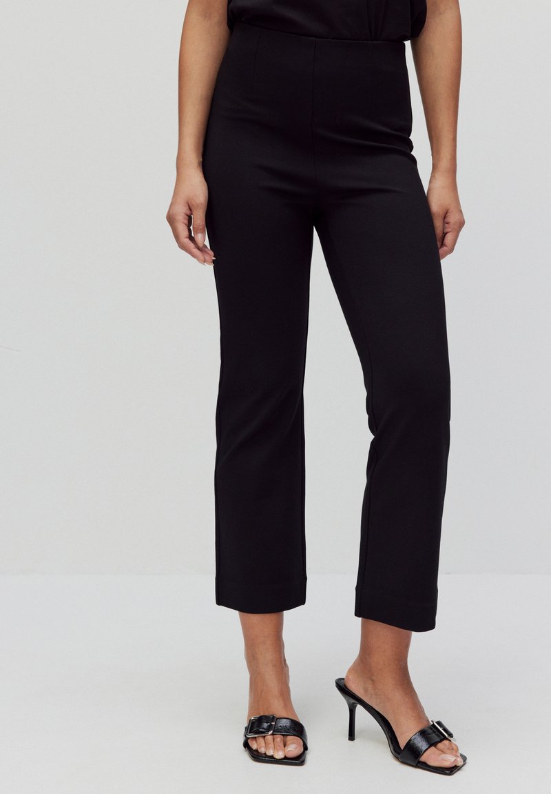 Woman wearing black cropped straight-leg pants and black heeled open-toe sandals with a buckle, standing against a plain background.