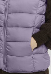 Lichtpaarse puffer vest met een gequilte uitstraling, voorzien van een rits aan de voorkant en zijzakken. Het model draagt een bruine gebreide trui eronder.