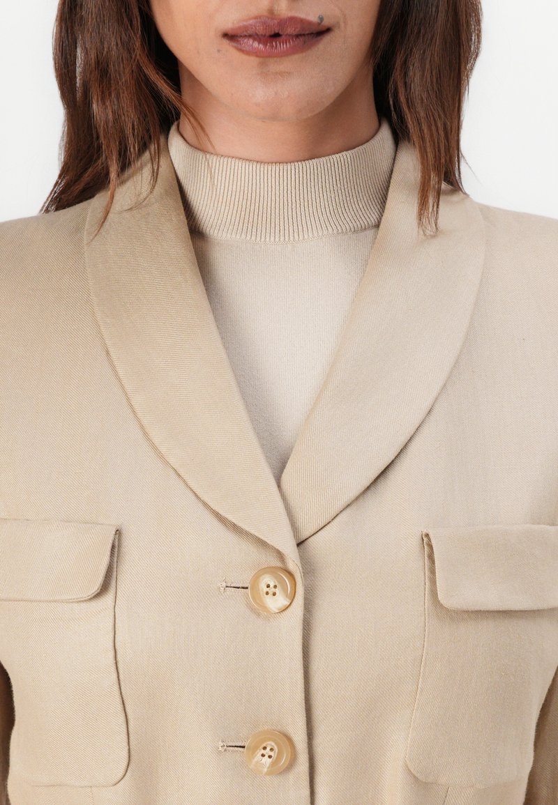 Beige Blazer mit zwei Taschen, Reverskragen und zwei Knöpfen. Texturiertes Material mit glatter Oberfläche. Über einem hellbeigen Rollkragenpullover getragen.