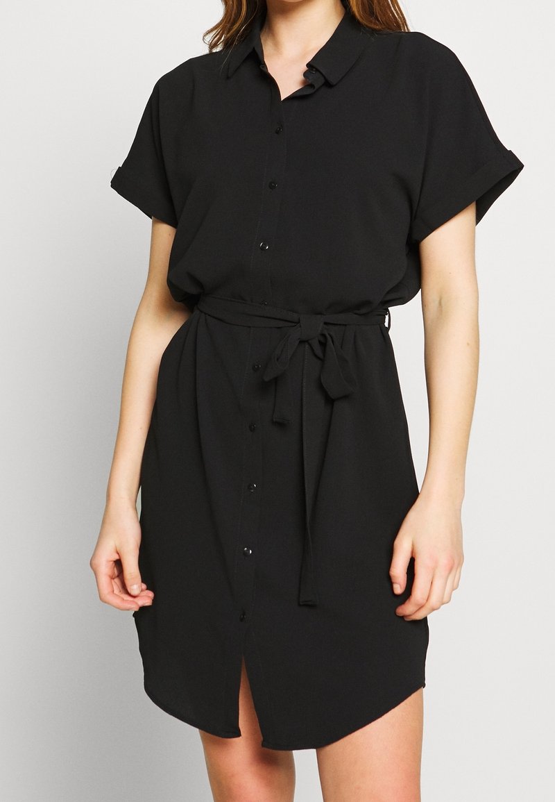 Vestido camisero negro hecho de tela ligera, con cuello tipo solapa, mangas cortas enrolladas, cierre frontal de botones y cintura ajustada con lazo.