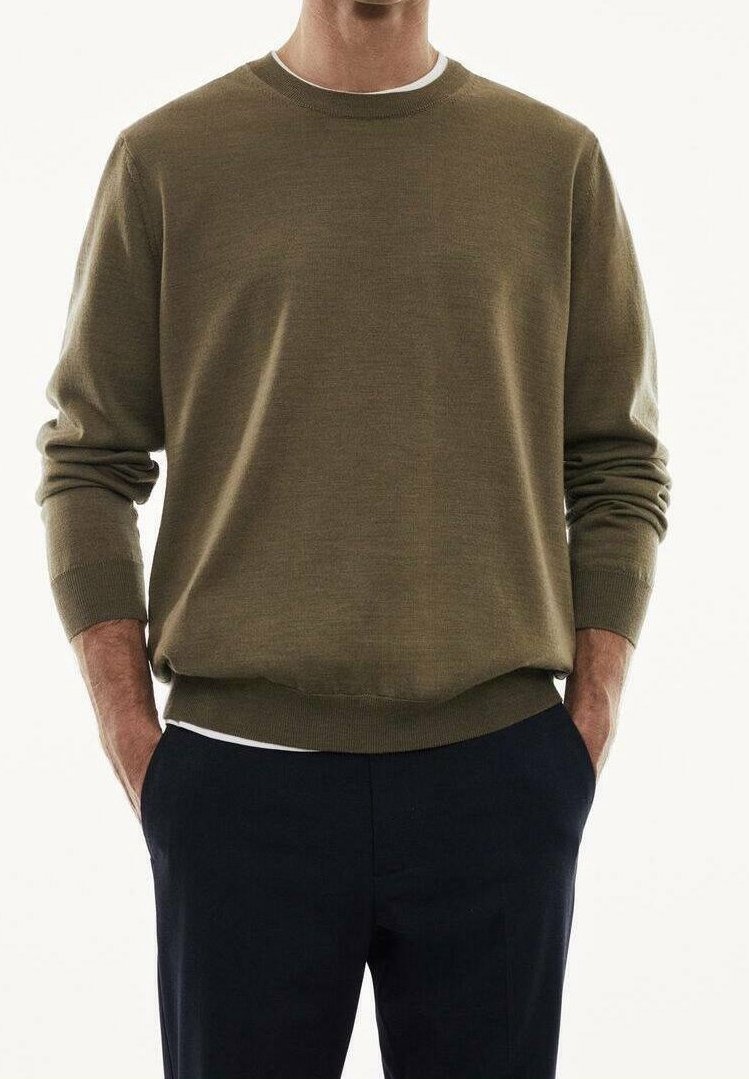 Homme portant un pull vert olive à manches longues et un pantalon noir, mains dans les poches, sur un fond blanc uni.