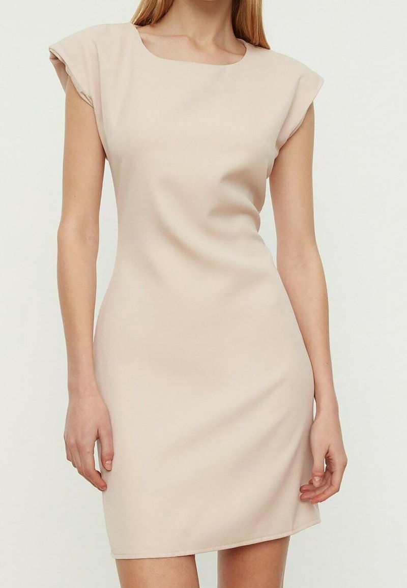 Femme portant une robe beige sans manches ajustée avec des épaules rembourrées, debout contre un fond clair uni.