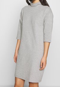 Šedé, kárované šaty po kolena vyrobené z měkké tkaniny s vysokým výstřihem a třičtvrtinovými rukávy. Jednoduchý, uvolněný střih bez dalších vzorů.