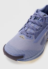 Scarpa sportiva viola con una tomaia in rete testurizzata, lacci rotondi e punta rinforzata. Presenta un logo e una suola in gomma a contrasto.