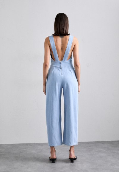 Jumpsuit en denim bleu clair avec des jambes larges, des bretelles réglables et un décolleté profond dans le dos. Comprend une fermeture éclair à l'arrière.