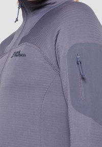 Primer plano de chaqueta Jack Wolfskin color lila claro con logotipo en el pecho, bolsillo con cremallera en el brazo y cuello alto con cordón ajustable.
