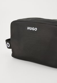 Fekete anyagú toilettás táska, téglalap alakú, egyik oldalán egy fehér "HUGO" logóval és azonos márkázású anyag fogantyúval.