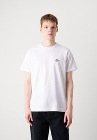 Hvid bomulds T-shirt med korte ærmer, rund halsudskæring, lille sort logo på venstre bryst og en afslappet pasform. Kombination med mørk denim jeans.
